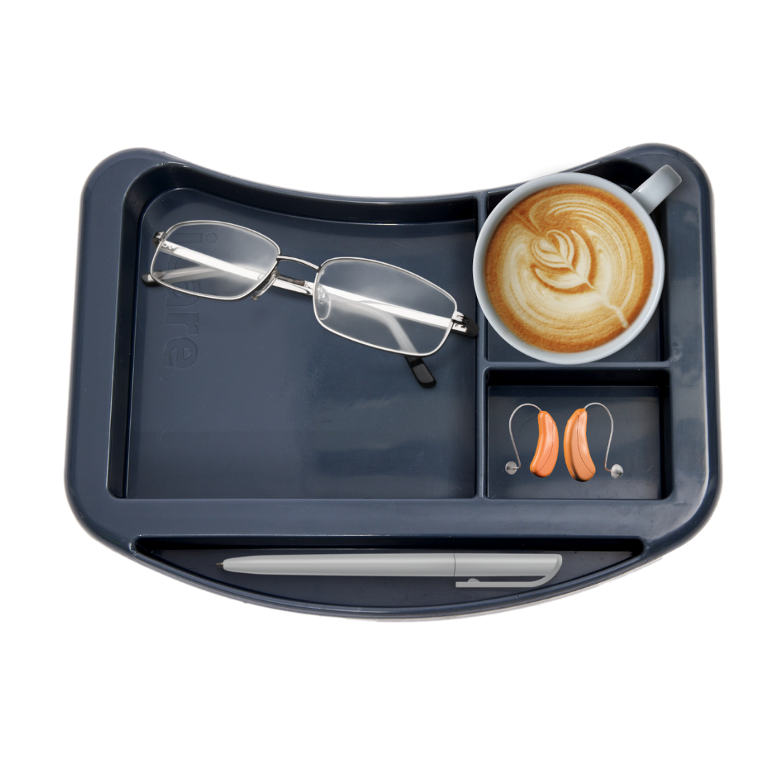 The iCare Medical Over Bed Tilting Table, in dark gray, serves as a versatile surface—holding glasses, a pen, cappuccino with latte art, and orange hearing aids.